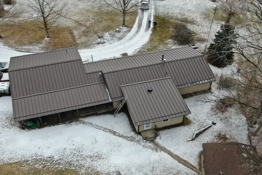 Brown standing seam metal roof under light snow, aerial view