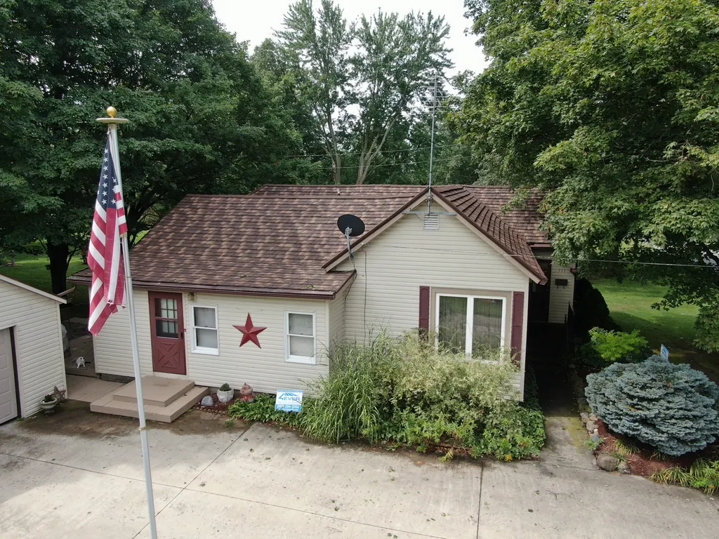 Small tan home with a reddish-brown stone-coated shake metal roof and an American flag